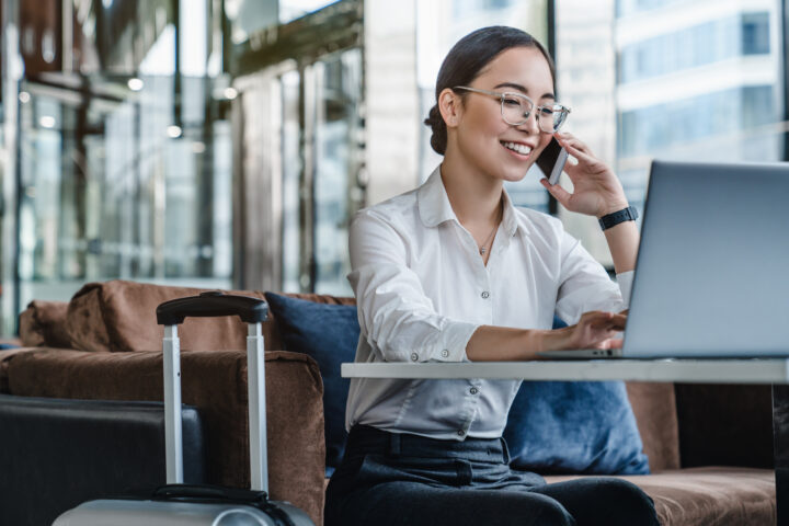 Asian female sitting at airport before trip having telephonic conversation with client and smiling.