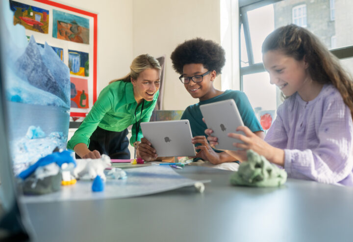 Teacher helping two students use tablet computers during a classroom art and science project, with artwork and model glaciers displayed on a table.