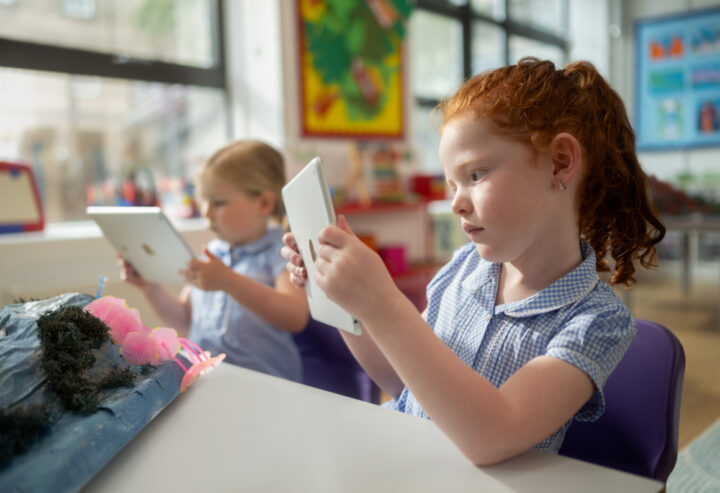 Two young students using tablet computers to examine a classroom project, sitting at a table with a handmade model display in a bright learning environment.