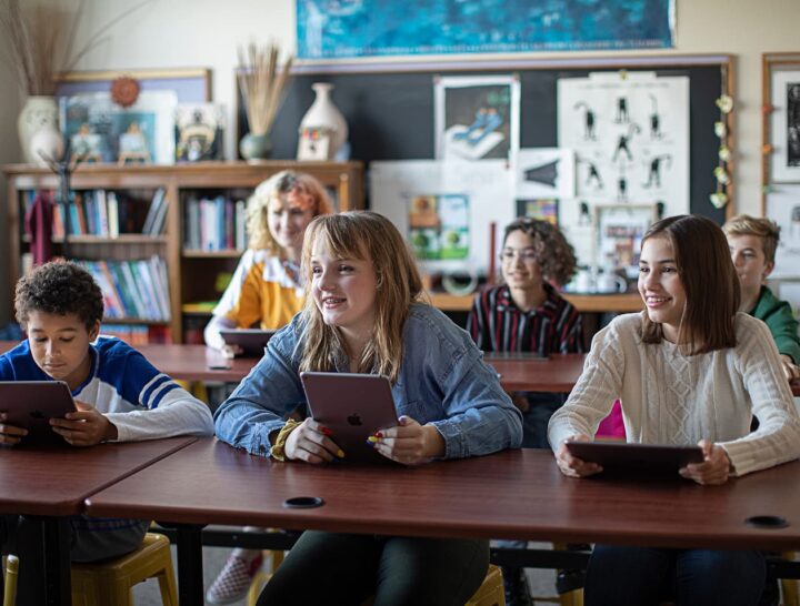 Students seated in a classroom using tablets during a lesson. Several students are engaged with their devices while participating in a collaborative and technology-enhanced learning environment.