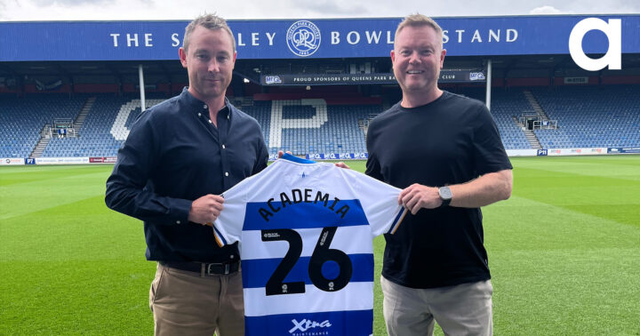 Two people stood on a football pitch holding a Queen Park Ranger's shirt with Academia and 26 on it.